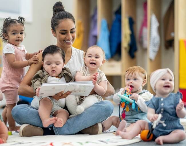 Trabajadora de guardería rodeada de bebés mientras está sentada en el suelo de la guardería.