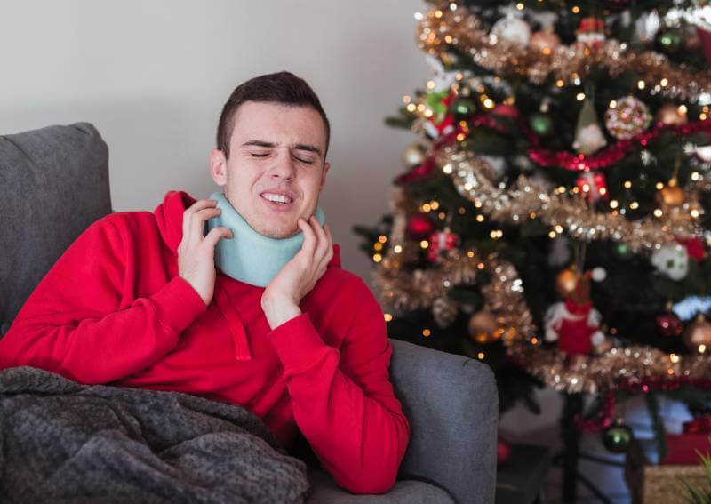 Joven con collarín sentado en un sofá junto a un árbol de Navidad.