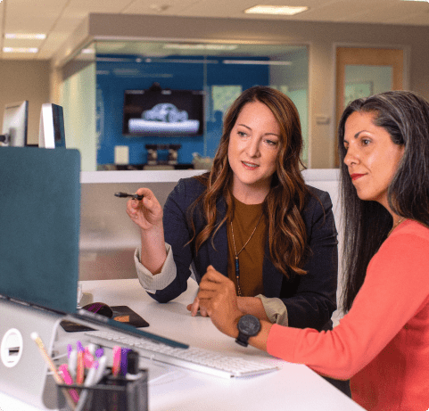 Dos mujeres en una oficina revisando información en el monitor de un ordenador.