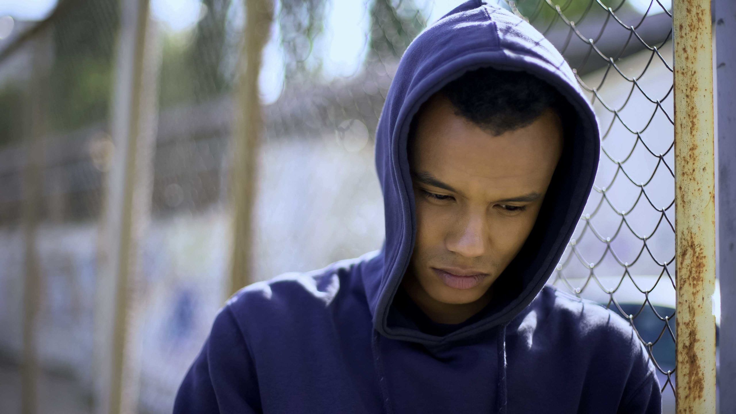 Teenager wearing a hoodie leaning against outside prison fence