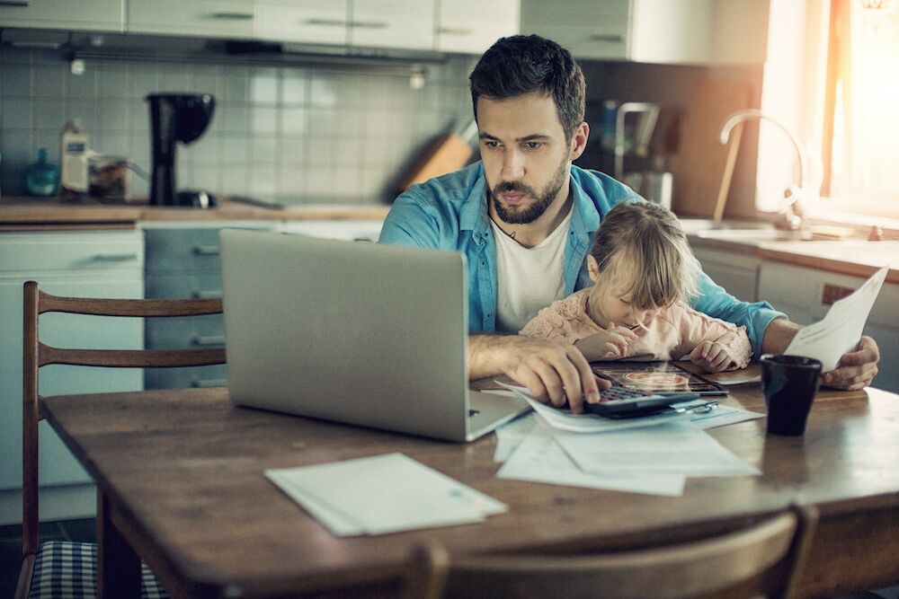 Father working at table in home kitchen with daughter on his lap playing a game