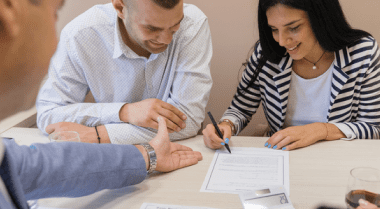 Woman signing a lease as her male partner and a landlord looks on.