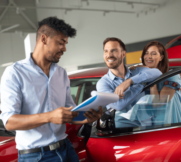 Happy couple negotiating a new car lease with a salesman inside an auto showroom. The car is red.