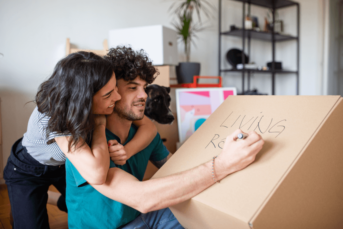 A man labeleling a moving box with Living Room as he girlfriend looks on.