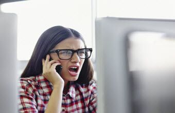 Young woman with angry expression talking on cell phone while looking at computer monitor