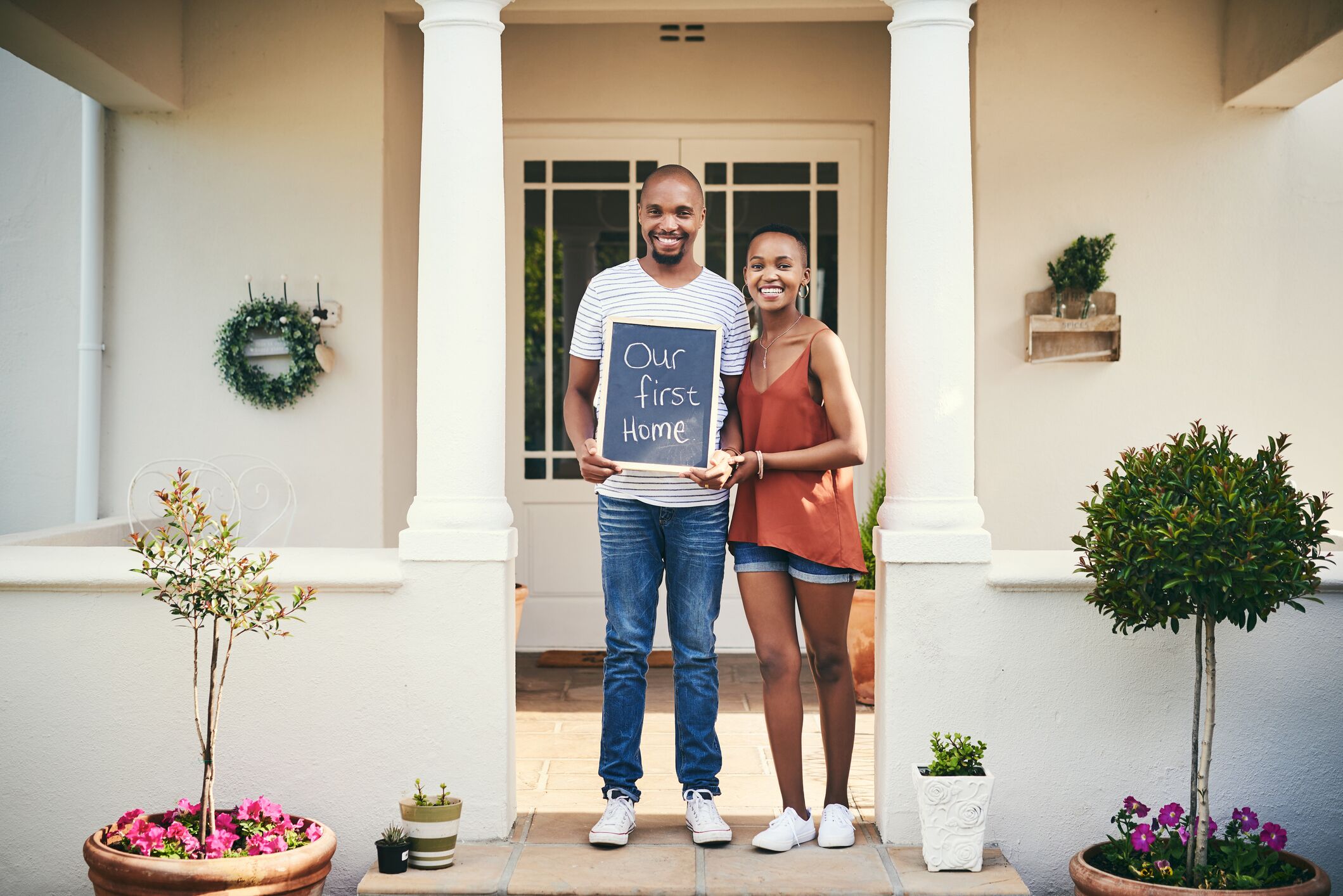 Pareja joven en las escaleras de su nueva casa con un cartel que dice: "Nuestra primera casa."