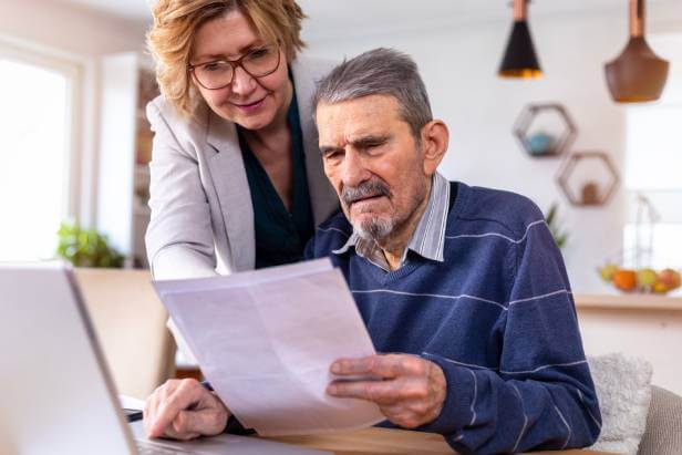 An elderly man is getting estate planning services from his lawyer as he reviews a legal document in front of a laptop. His lawyer is standing beside him as he sits at a table.