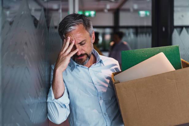 Distraught man recently dismissed from his job. He's holding a box of containing his office belongs and leaning against a workplace wall.