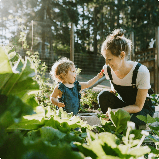 Una madre trabaja en su jardín mientras su hija pequeña le da un pétalo de flor.