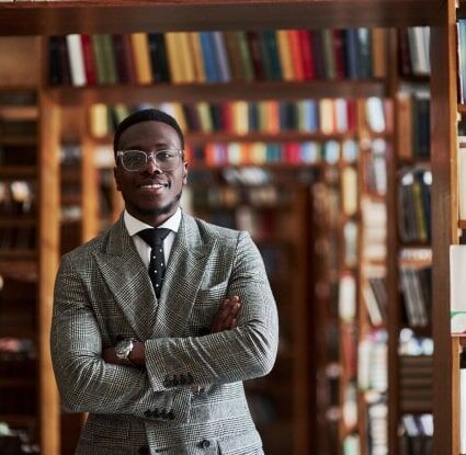 Un abogado especializado en propiedad intelectual de pie en una biblioteca jurídica con los brazos cruzados y sonriendo.
