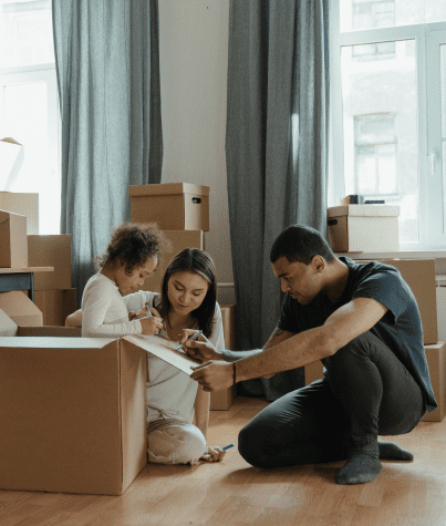 Una madre, un padre y una niña etiquetan cajas de mudanza en el salón de su casa.