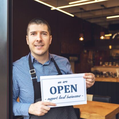 Un empresario en la puerta de su restaurante con un cartel en el que se puede leer: estamos ABIERTOS. apoye a los negocios locales.