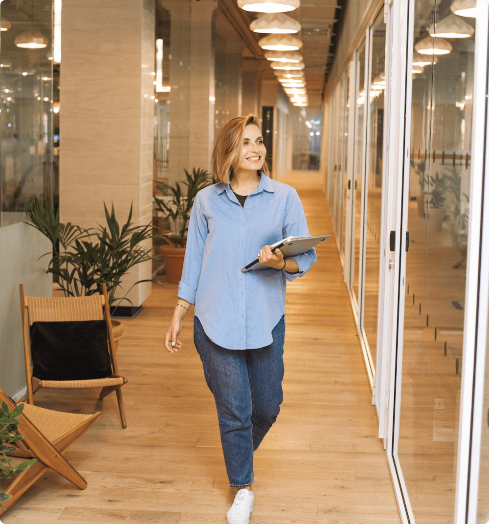 Una empresaria sonriente caminando por el pasillo de su oficina con un ordenador portátil.