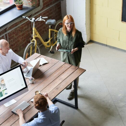 Un empresario de pie junto a una mesa de oficina sosteniendo un ordenador tablet mientras dos empleados varones trabajan en una mesa cercana.