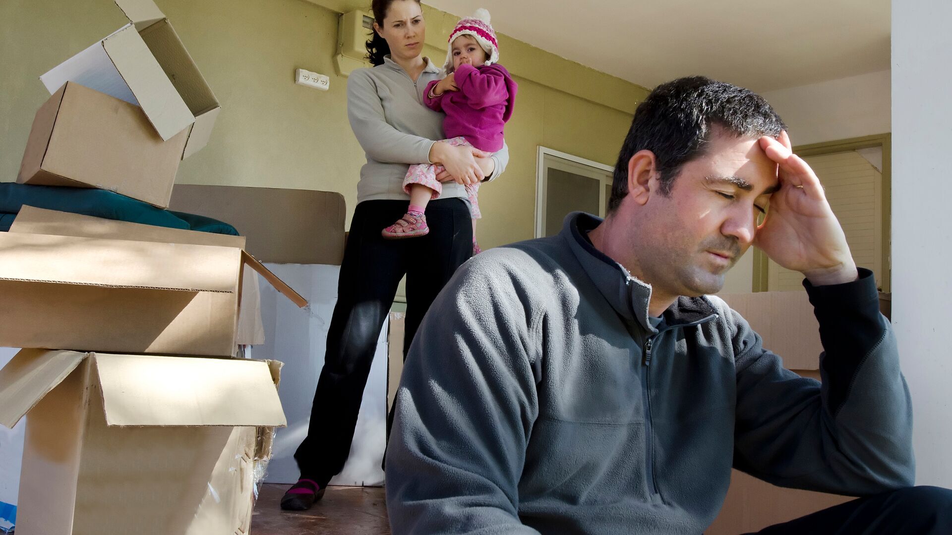 Stressed man sitting down as a woman holding a young child look on in a house being packed for a move.
