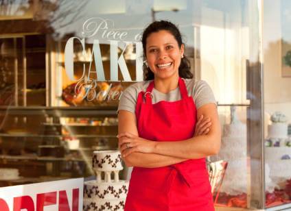 Smiling woman in a red apron in front of a bakery she owns who has learned how to create a DBA for her LLC.