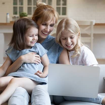 Una madre sonriente y sus dos hijas pequeñas miran la pantalla de un ordenador portátil sentadas en el sofá.