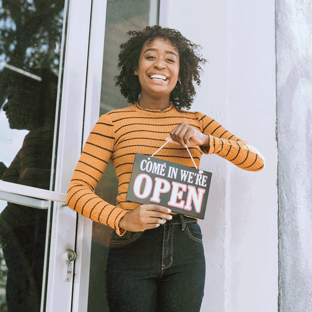 LegalShield Tips for Small Business Week 2022: Happy young business owner holding a sign that reads, Come In We're Open
