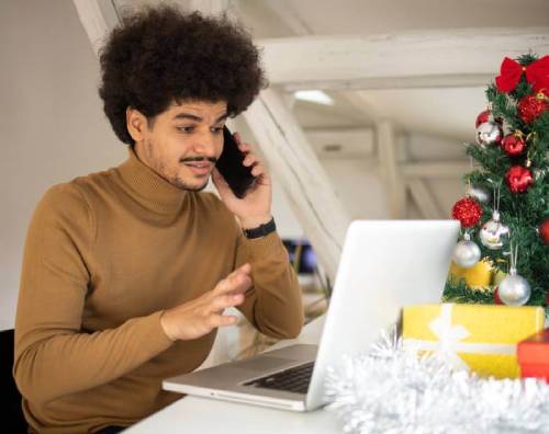 Stressed man talking on his phone while looking at his laptop as he sits near a Christmas tree.